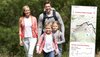 Eine Familie mit zwei Kindern wandert durch eine grüne Landschaft. Rechts vorne ist ein Ausschnitt einer App zu sehen.
