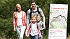 Eine Familie mit zwei Kindern wandert durch eine grüne Landschaft. Rechts vorne ist ein Ausschnitt einer App zu sehen.