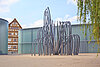 Moderne Metallskulptur vor einem Fachwerkhaus und einem modernen Gebäude.