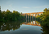 Eisenbahnbrücke über ruhigen See mit Spiegelung, umgeben von grünen Bäumen.