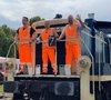 Drei Lokführer in orangen Warnwesten und -hosen winken fröhlich von Aufstieg eines Triebfahrzeugs.