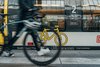 Im Vordergrund ist verschwommen eine Person zu erkennen, die ihr Fahrrad das Bahngleis entlangschiebt. Im Hintergrund sieht man eine Bahn mit einem Fahrradsymbol an der Seite.
