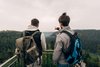 Zwei Männer mit Outdoor-Kleidung und großen Rucksäcken stehen mit dem Rücken zur Kamera an einem Aussichtspunkt mit Geländer. Einer zeigt mit dem Arm auf das bewaldete Tal vor ihnen. Die Landschaft ist hügelig, der Himmel ist bewölkt.