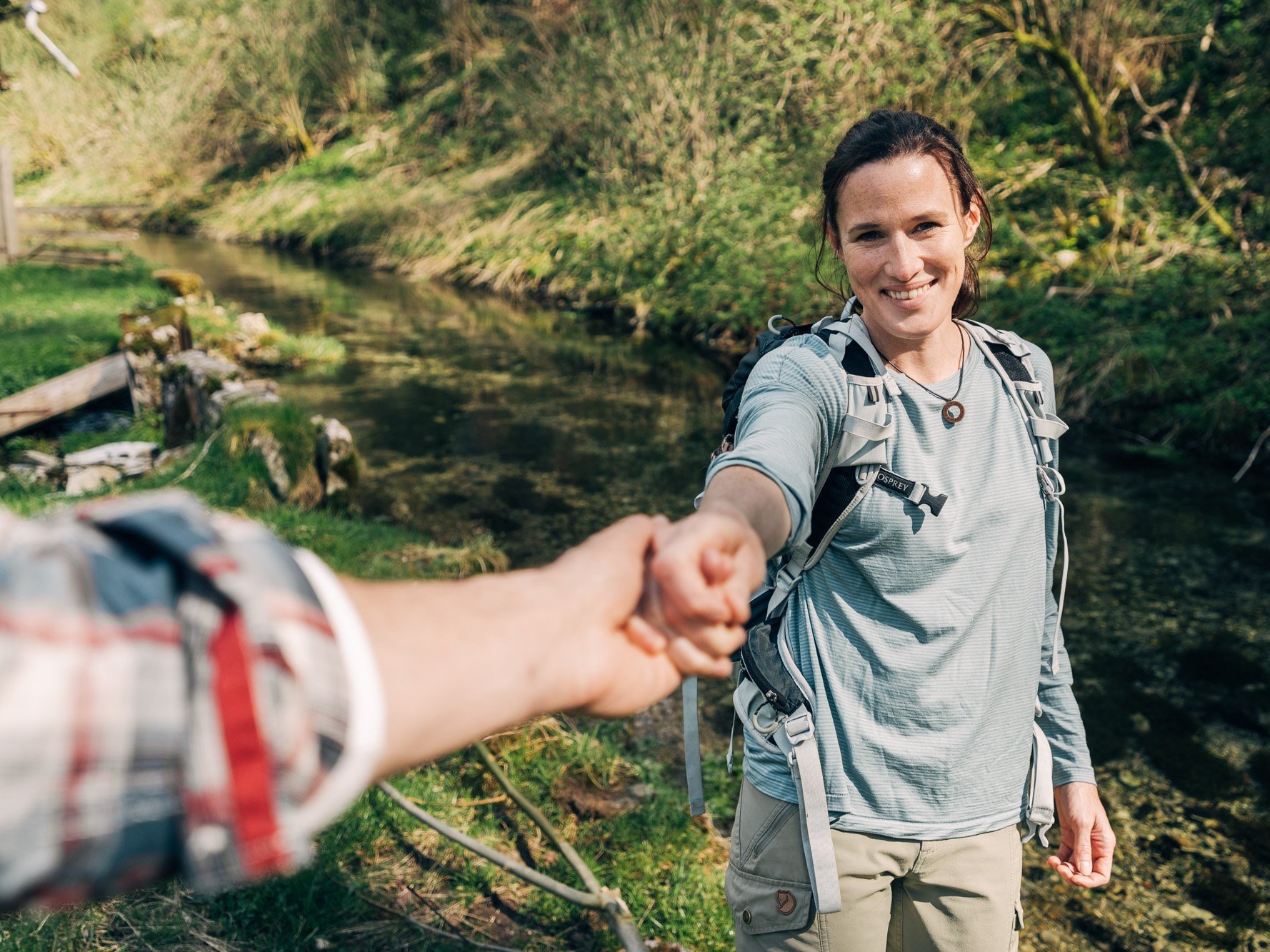 Eine Hand ragt ins Bild und greift die Hand einer Frau in Wander-Klamotten. Die Frau blick zurück in die Kamera.