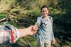 Eine Frau mit Outdoor-Kleidung und Rucksack steht lächelnd am Rand eines klaren Bachs in einer bewaldeten Landschaft. Sie streckt dem oder der Fotograf*in die Hand entgegen. Nur ein Arm im karierten Hemd ist im Vordergrund zu sehen.