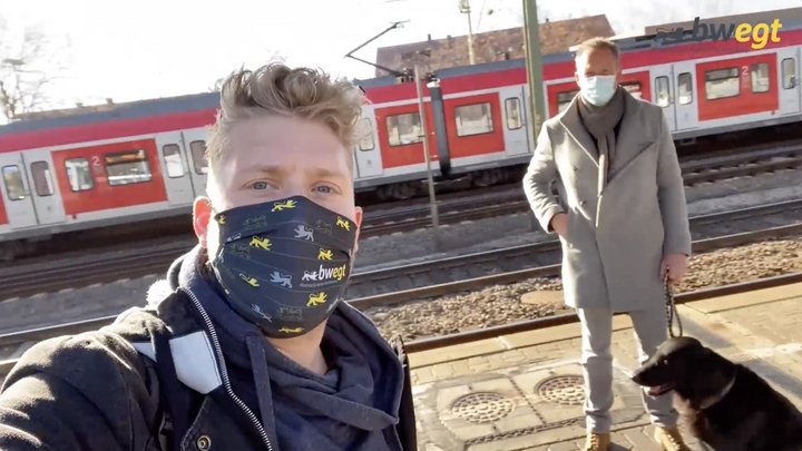 Mobilitäts-Checker Kevin steht maskiert am Bahnsteig. Im Hintergrund steht ein weiterer maskierter Mann mit Hund an der Leine.