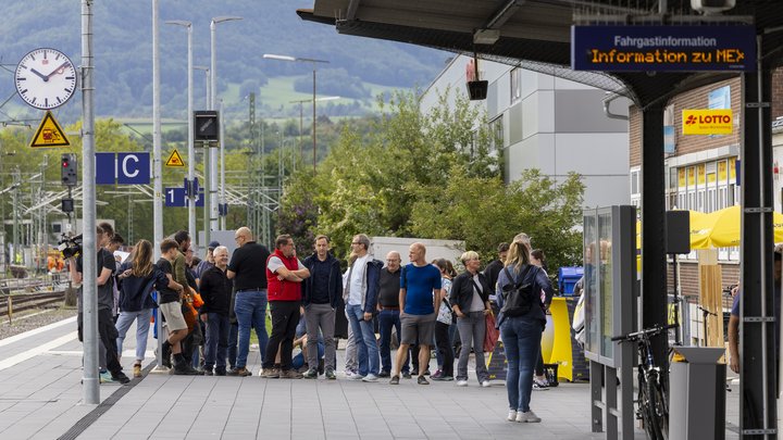 Eine Gruppe von Menschen steht an einem Bahnhof, im Hintergrund ist ein hügeliger Wald zu sehen