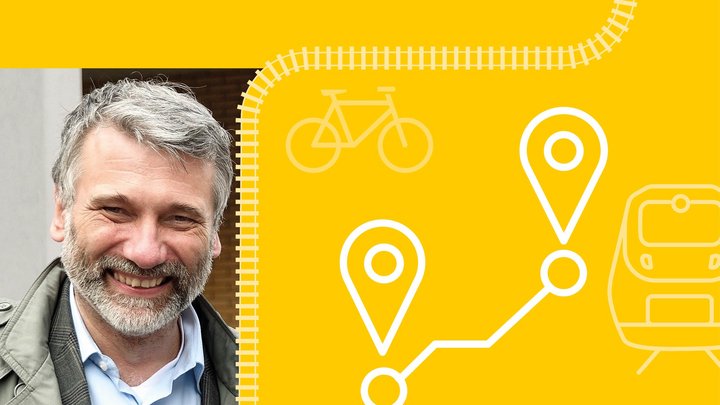 Ein Mann mittleren Alters mit grauen Haaren und grauem Bart lächelt in die Kamera. Er trägt ein Hemd und eine offene Jacke.