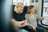 Mittelalter Herr mit weißen Haaren und Bart sitzt neben einer mittelalten Frau mit brauen Haaren im Zug. Die Frau schaut auf ihre Uhr am Handgelenk, der Mann verschränkt seine Arme.
