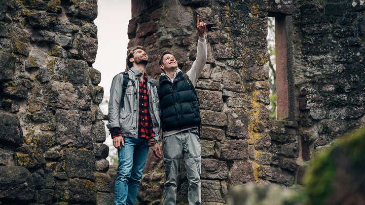 Zwei junge Männer stehen in einer Ruine und blicken nach oben. Ein Mann zeigt auf etwas.