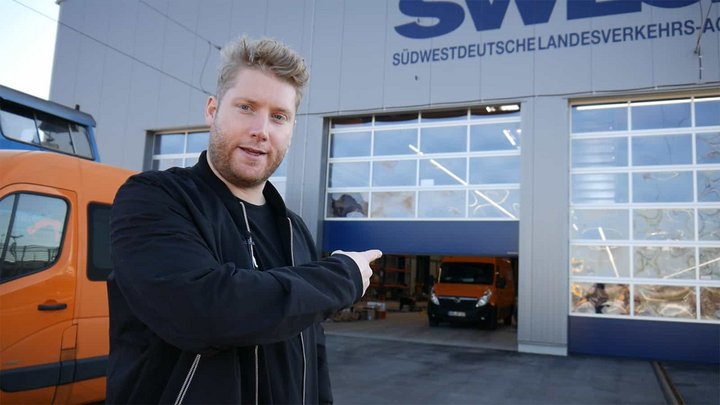 Mann steht vor einer Bahnbetriebswerkstatt und zeigt auf ein sich öffnendes Tor.