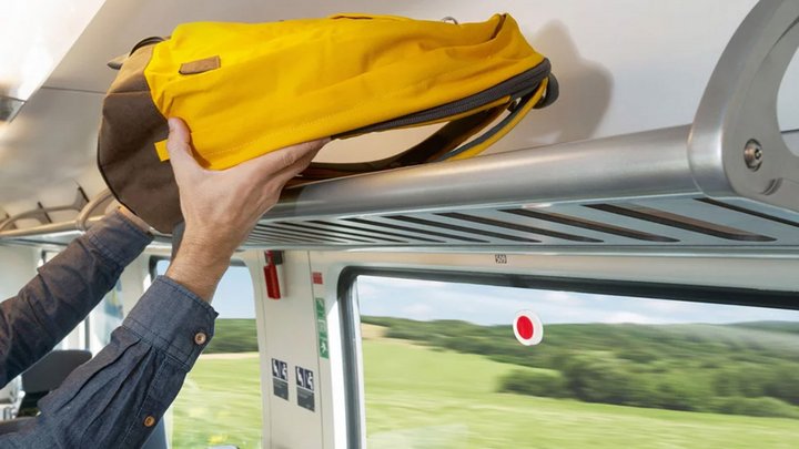 Ein Mann verstaut seinen gelben Rucksack im Zug auf einer Ablage