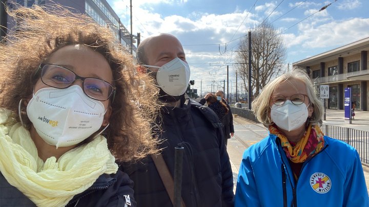 Blinder Mann steht mit zwei Frauen am Bahnhof.