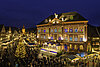 An einem Platz mit Weihnachtsmarkt und großem Tannenbaum steht ein mit Leuchten angestrahltes Rathaus. Die Fenster schmücken kunstvolle Motive.