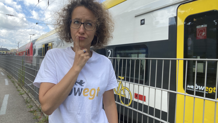 Frau steht vor einem Zug und hält nachdenklich einen Finger an die Lippen.