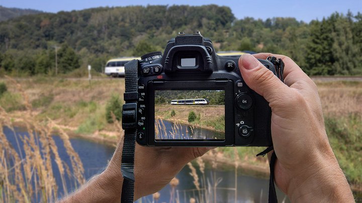 Eine Kamera fotografiert einen bwegt-Zug in einer Landschaft mit Fluss und Wald.