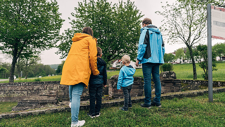 Familie mit zwei kleinen Kindern steht mit dem Rücken zur Kamera vor den Überbleibseln einer römischen Ruine.
