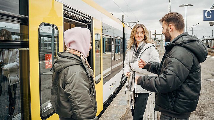 Drei junge Menschen sind mitten im Gespräch, während sie in eine Bahn einsteigen.