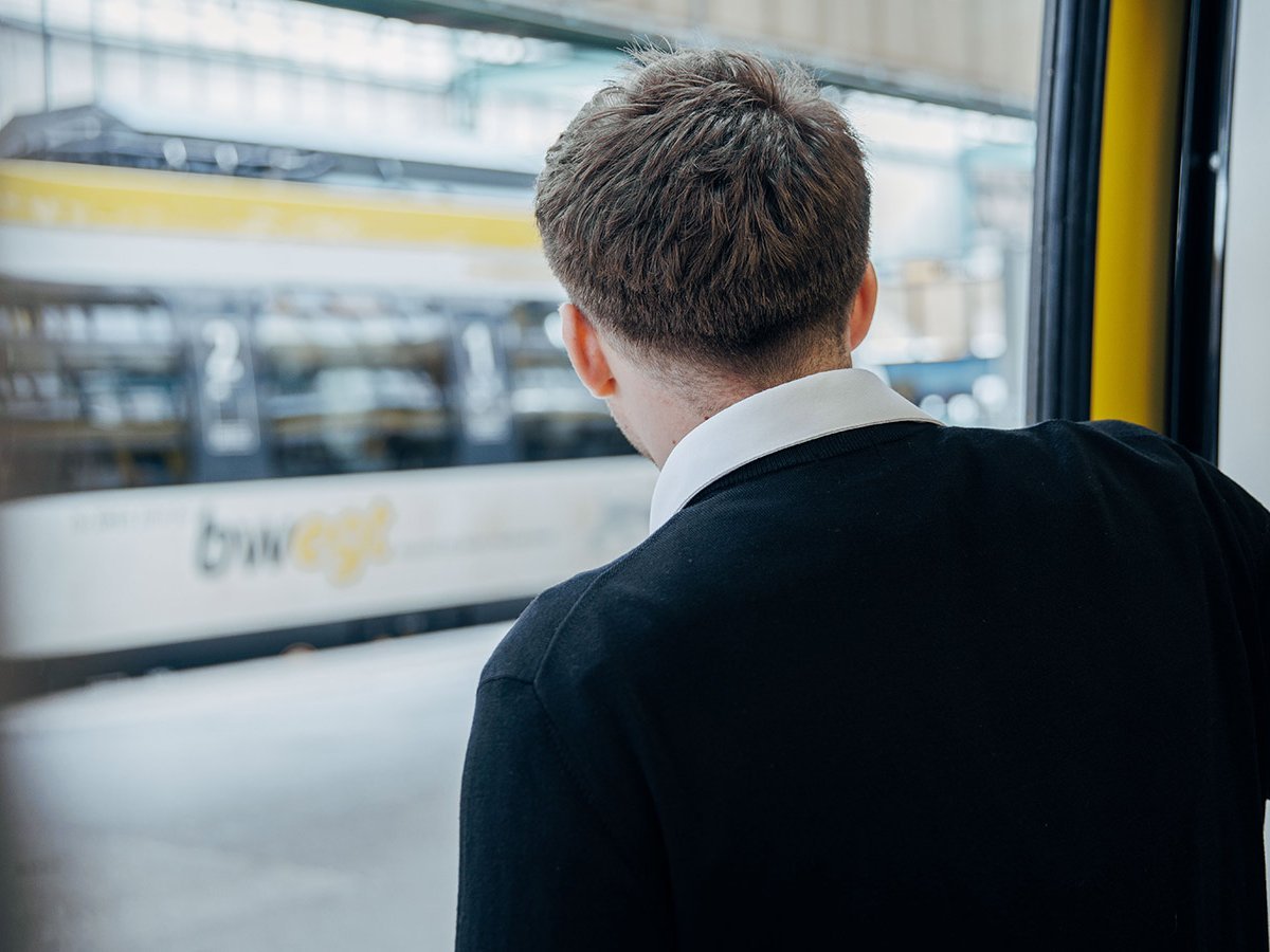 Mit dem Rücken zur Kamera gedrehter Mann lehnt sich aus einem Zug auf den Bahnsteig. Dort steht im Hintergrund noch ein verschwommen sichtbarer Zug.