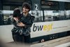 Ein junger Mann mit lockigen dunklen Haaren umarmt eine Frau in schwarzer Lederjacke vor einem stehenden bwegt-Zug. Beide lächeln sich an. Auf dem Zug ist deutlich das bwegt-Logo mit dem Schriftzug „Mobilität für Baden-Württemberg“ sichtbar.