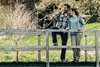 Ein Paar steht auf einer alten Holzbrücke in grüner Umgebung. Der Mann hält eine Kamera hoch fürein Foto das Gesicht zur Kamera gewandt. Die Frau lehnt sich an ihn, beide tragen Outdoor-Kleidung und Rucksäcke.
