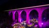 Bild bei Nacht einer violett angestrahlten Bahnbrücke. Darunter im Tal sind die Hütten eines Weihnachtsmarkts zu sehen.