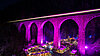 Bild bei Nacht einer violett angestrahlten Bahnbrücke. Darunter im Tal sind die Hütten eines Weihnachtsmarkts zu sehen.