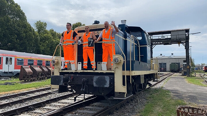 Drei Auszubildende Lokführer stehen auf einer Bahn und winken.