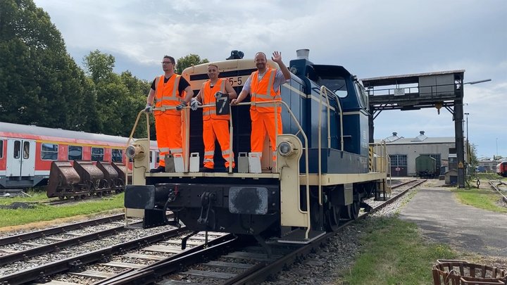 Drei Auszubildende Lokführer stehen auf einer Bahn und winken.