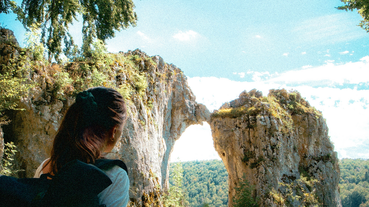 Frau mit Wanderrucksack steht vor der Felsformation „Küssende Sau“ in Blaubeuren, der Himmel ist blau, am Horizont ist ein Wald zu erkennen