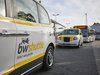 Zwei London Taxis als Shuttles im gelb-weiß-grauen Design. Auf dem Fahrzeug im Vordergrund ist die Aufschrift „bwshuttle“ an der Seite im Fokus. Sie stehen hintereinander seitlich an einer Haltestelle. Dahinter ein Linienbus.