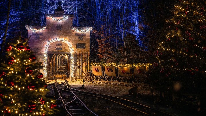 Eingang eines Bergwerks verziert mit Lichterketten und Schmuck, die am Abend leuchten. Auch die Tannen und Wagen drumherum sind geschmückt.