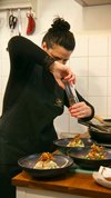  Eine Restaurant-Köchin mit in schwarzer Kleidung und Schürze mahlt mit einer Pfeffermühle über drei filigran angerichtete Teller.