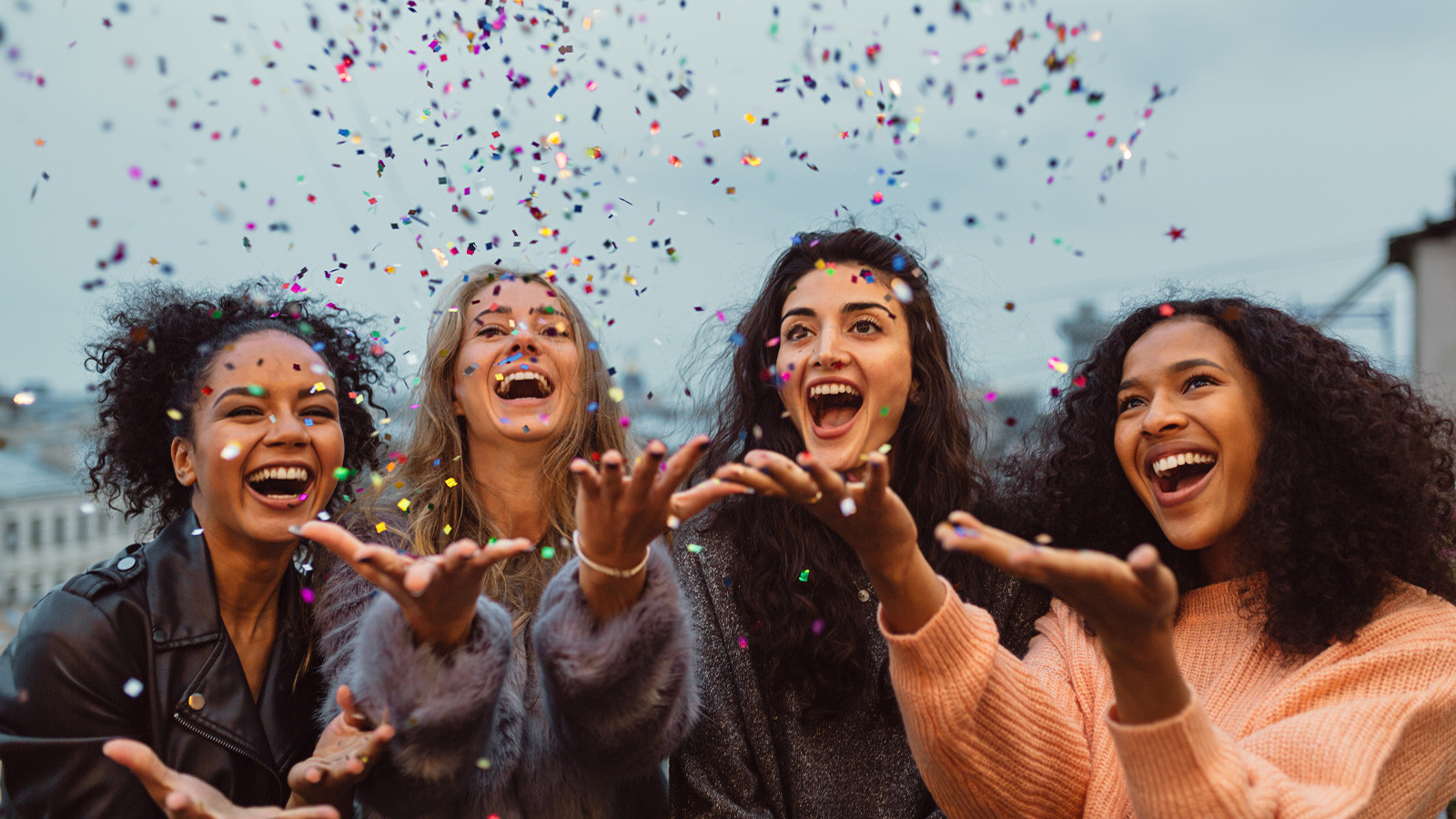 Vier junge Frauen lachen auf einer Veranstaltung und werfen Konfetti hoch.