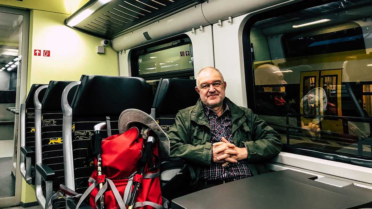 Mann sitzt in bwegt-Zug und neben ihm auf dem Sitz steht ein roter Wanderrucksack.