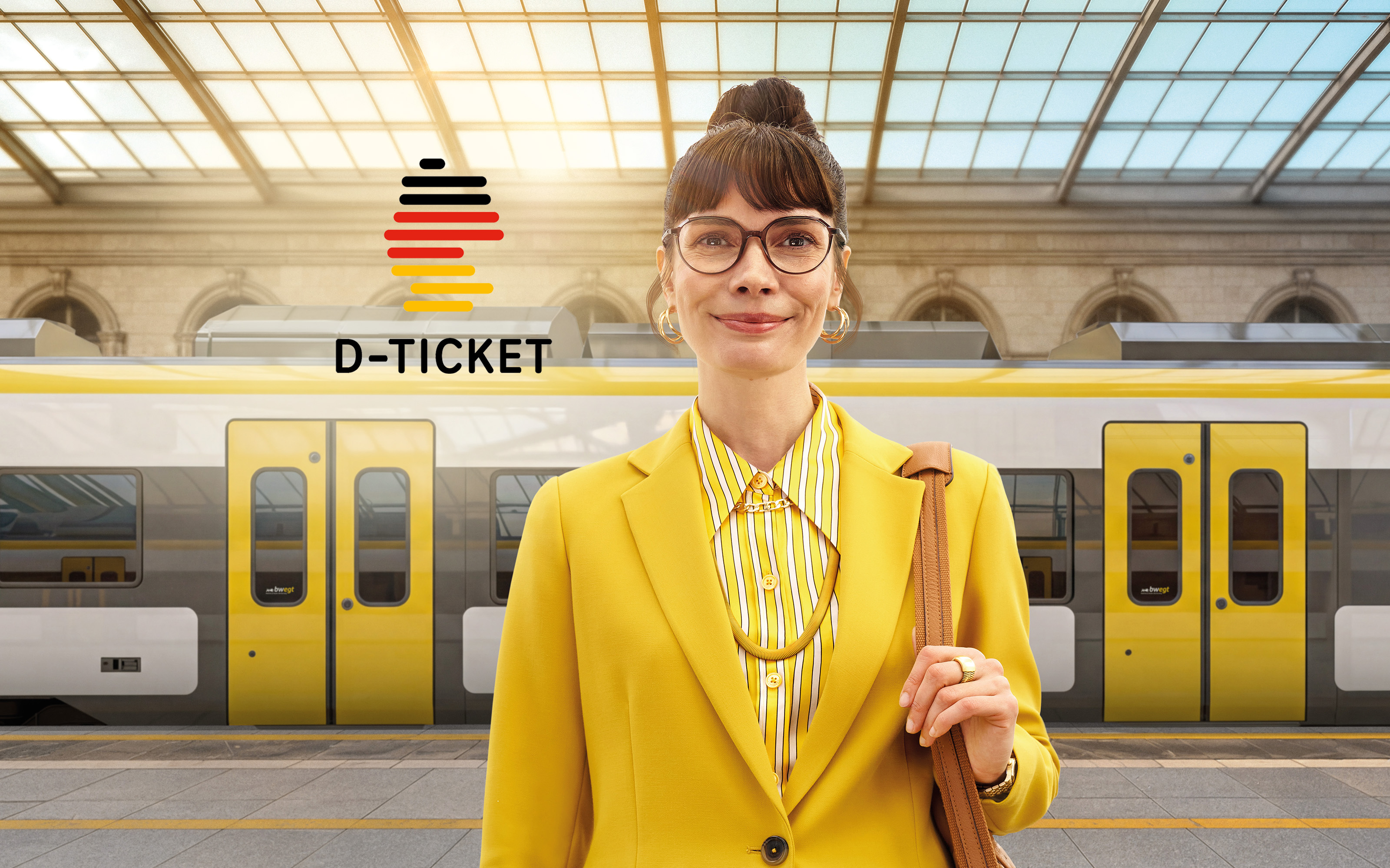 Eine Frau in gelber Kleidung steht an einem Bahnhof. Hinter ihr fährt ein bwegt-Zug vorbei.