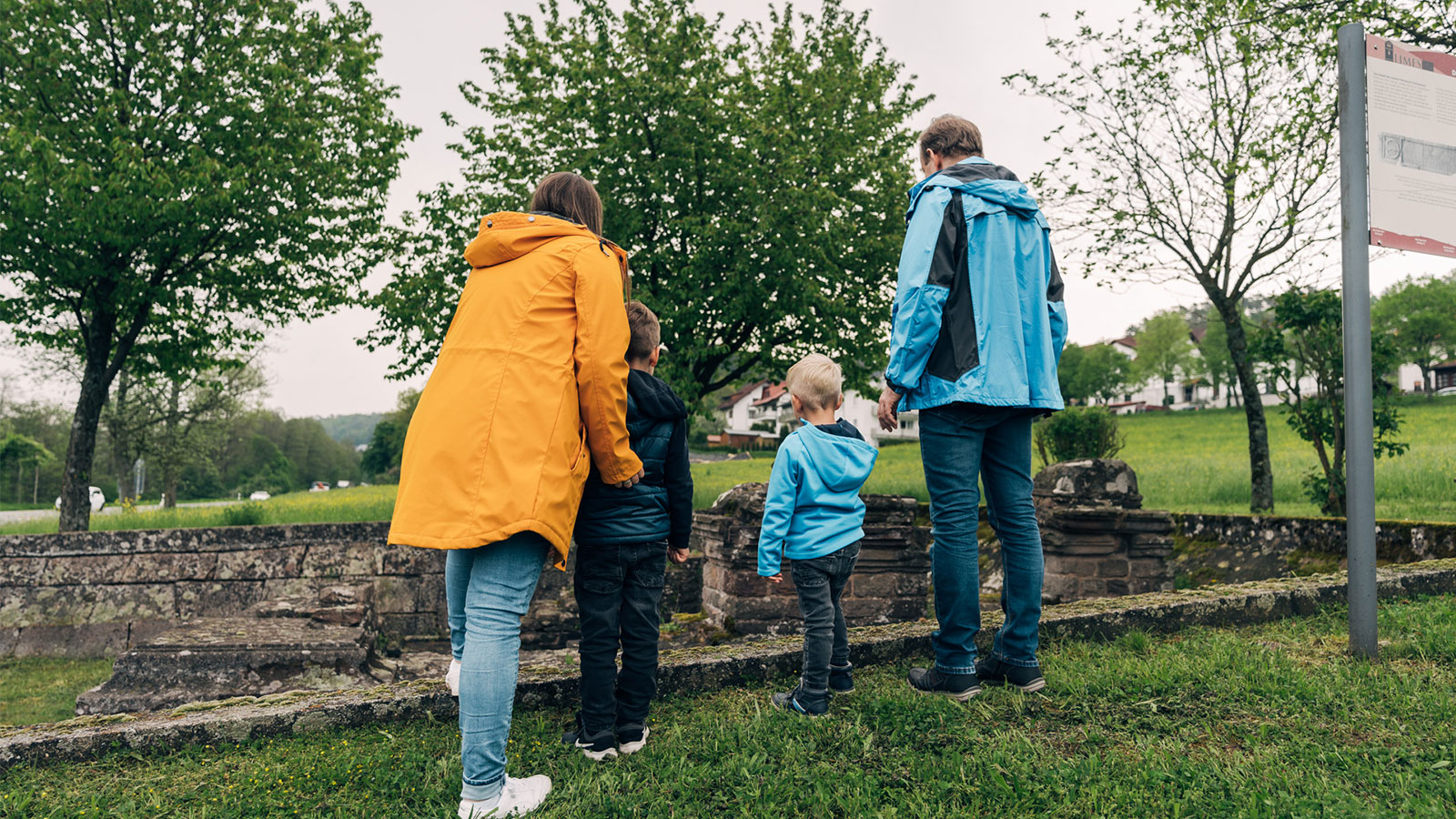 Eine Familie mit zwei Kindern steht mit dem Rücken zur Kamera vor den Überbleibseln einer römischen Ruine.