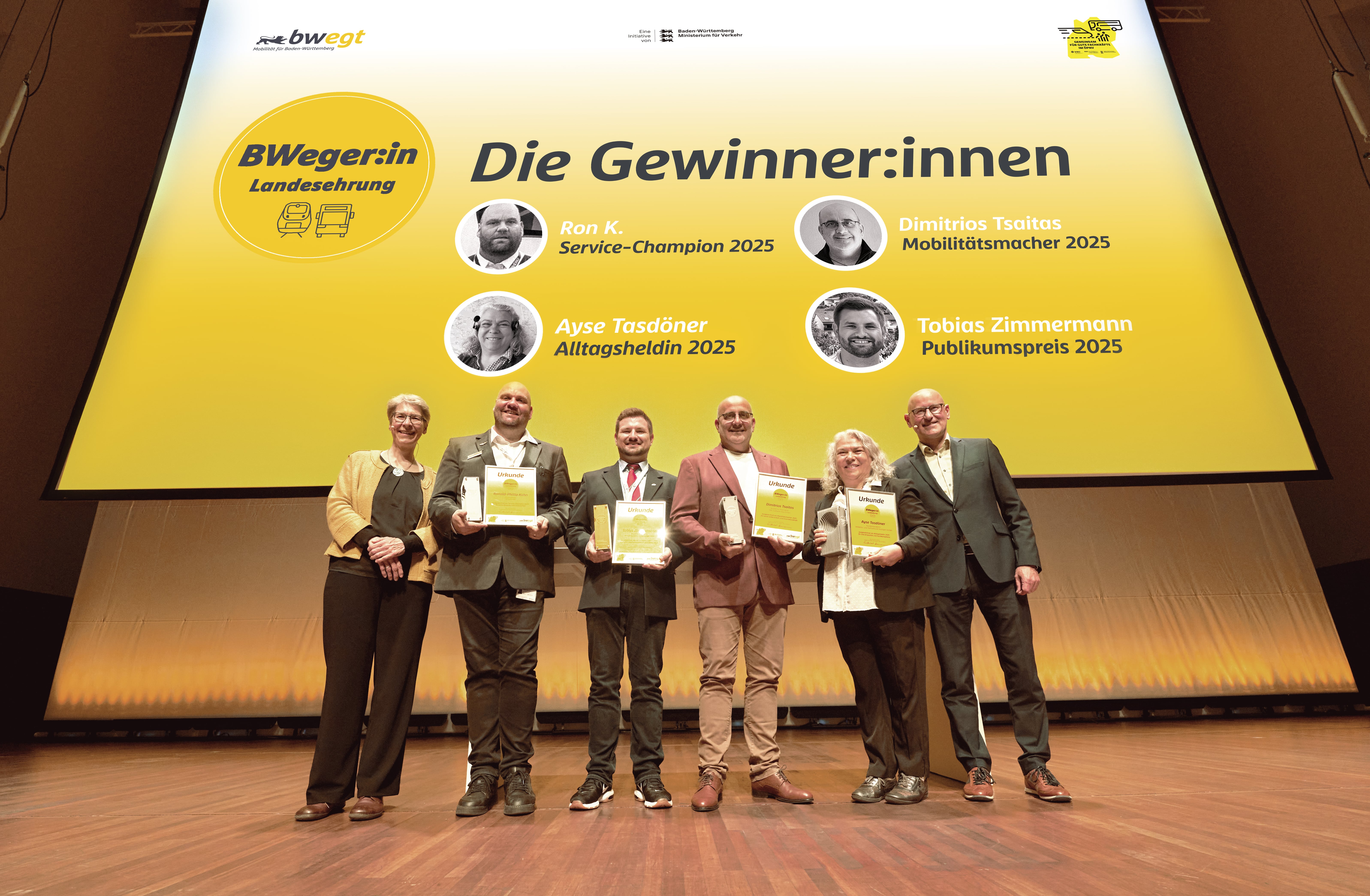 Auf einer Bühne präsentieren vier Preisträger:innen in festlicher Kleidung ihre Siegerurkunden und Trophäen. Die Aufschrift im Hintergrund lautet: „BWeger:in Landesehrung. Die Gewinner:innen: Ron K. Service-Champion 2025, Ayse Tasdöner Alltagsheldin 2025, Dimitrios Tsaitas Mobilitätsmacher 2025, Tobias Zimmermann Publikumspreis 2025“. 