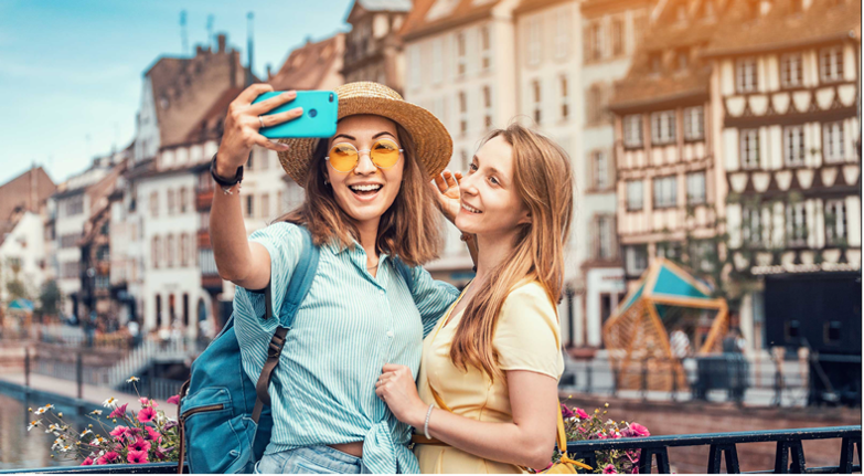 Zwei junge Frauen stehen vor alten Fachwerkhäusern in Frankreich und machen ein Selfie. 