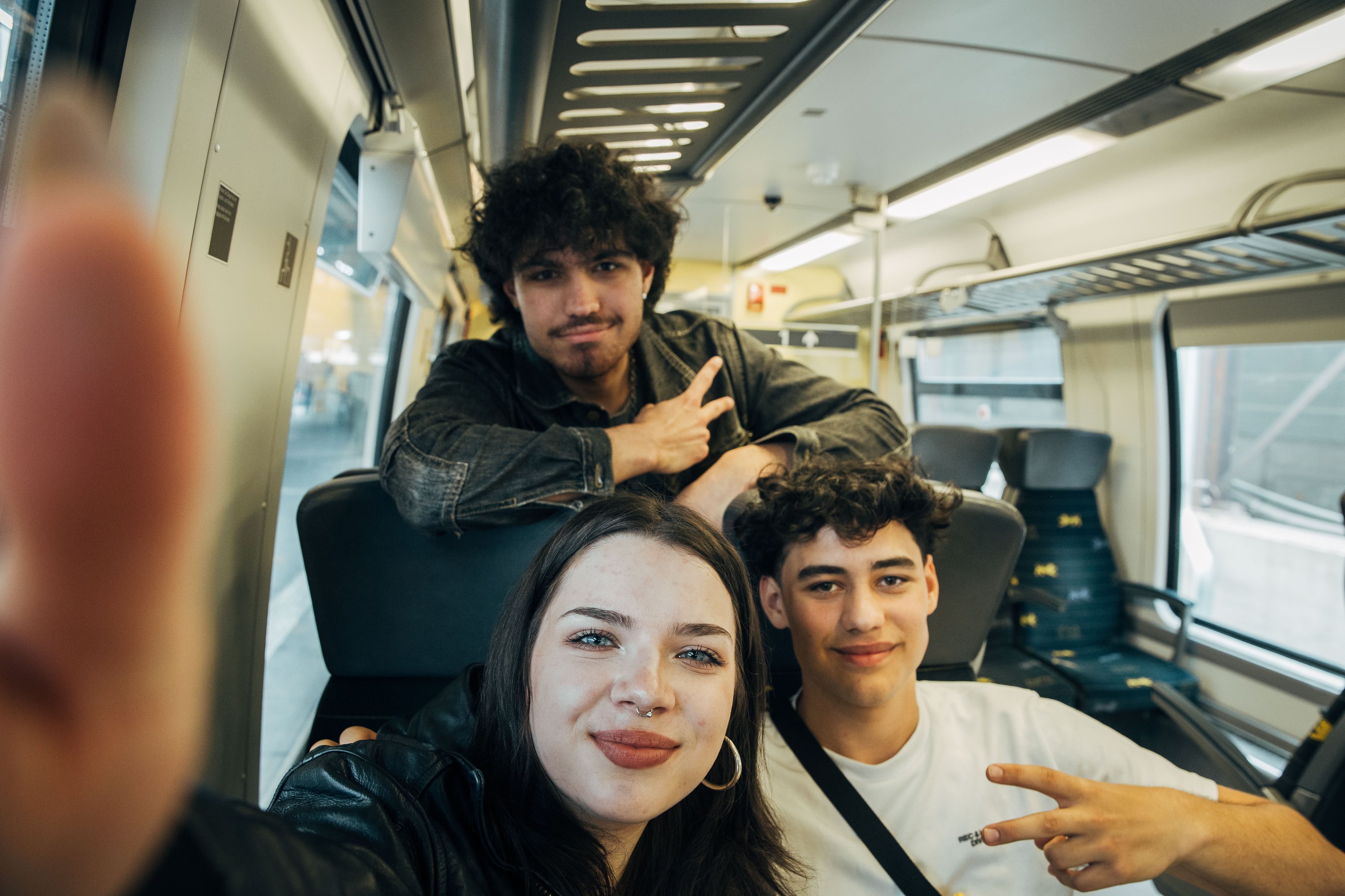 Drei Jugendliche sitzen zusammen in der Bahn und machen ein Selfie. Die beiden Jungen zweigen ein Peace-Zeichen, das Mädchen hält das Smartphone.