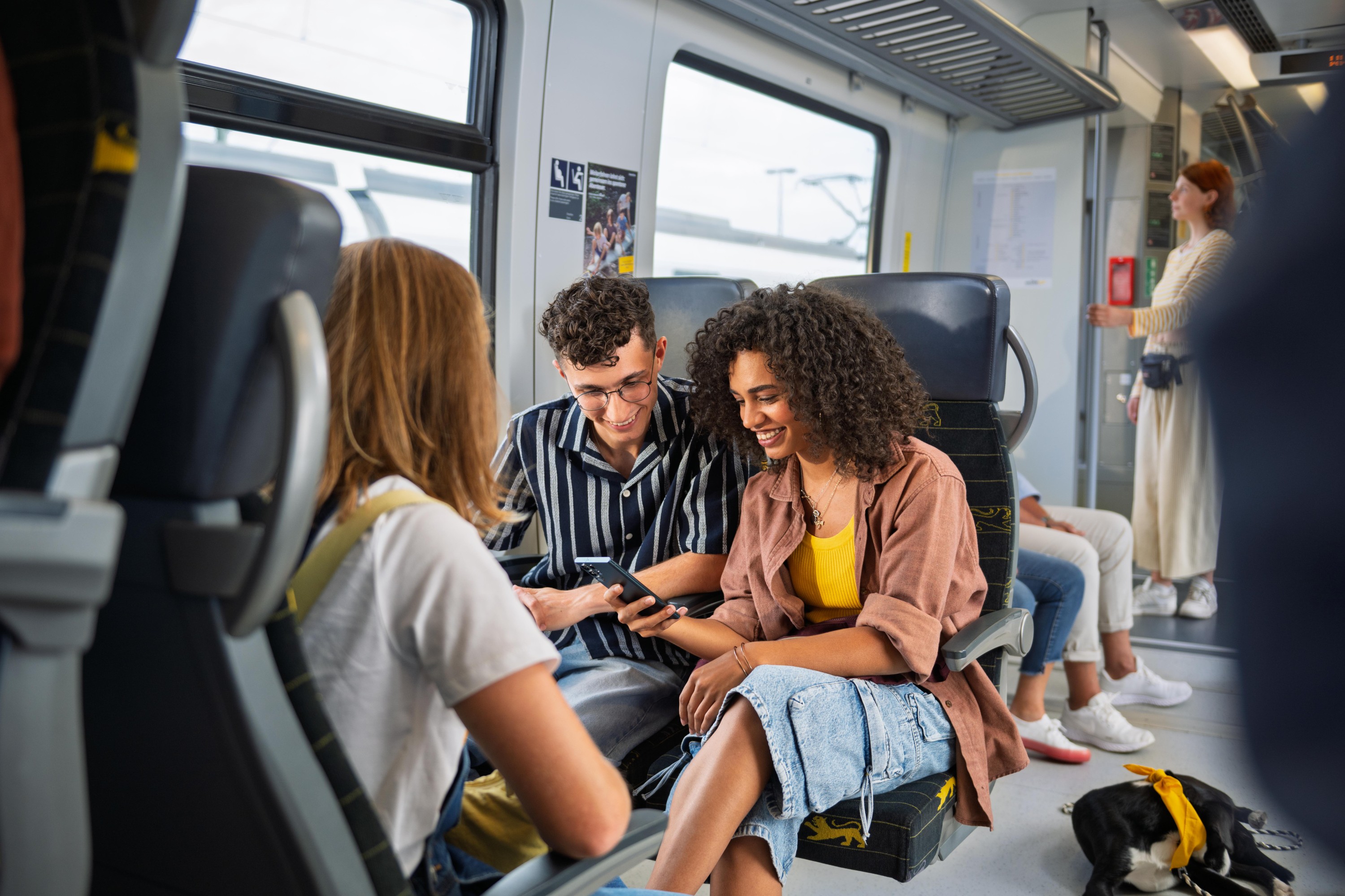 Eine Gruppe Jugendlicher sitzt zusammen in einem Vierer in der Bahn. Die zwei der Kamera zugekehrten Personen schauen lachend auf ein Smartphone.