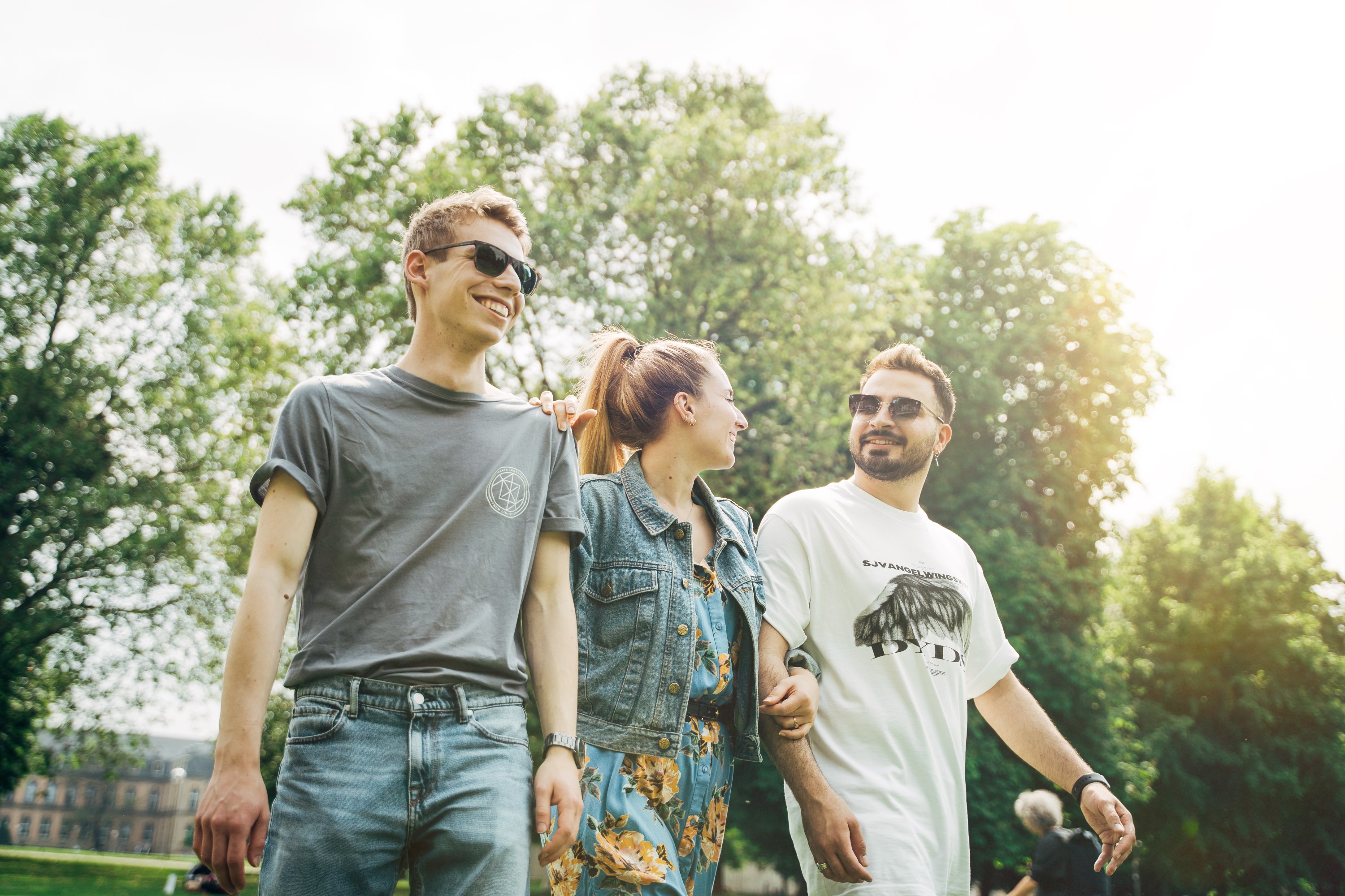 Drei junge Leute gehen lachend und sich in den Armen liegend durch einen Park. Es scheint die Sonne und sie sind sommerlich angezogen.