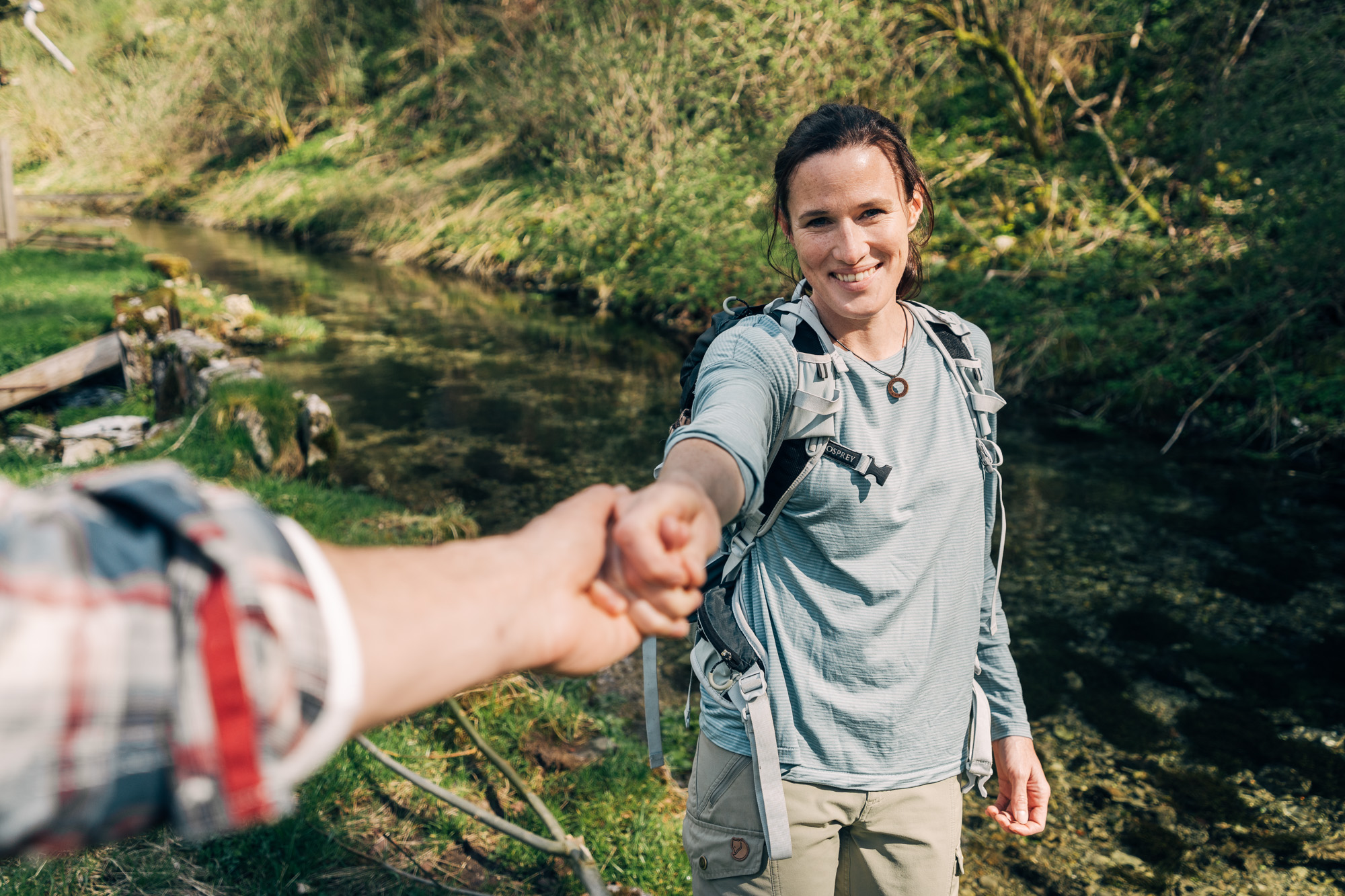 Eine Frau mit Outdoor-Kleidung und Rucksack steht lächelnd am Rand eines klaren Bachs in einer bewaldeten Landschaft. Sie streckt dem oder der Fotograf*in die Hand entgegen. Nur ein Arm im karierten Hemd ist im Vordergrund zu sehen.