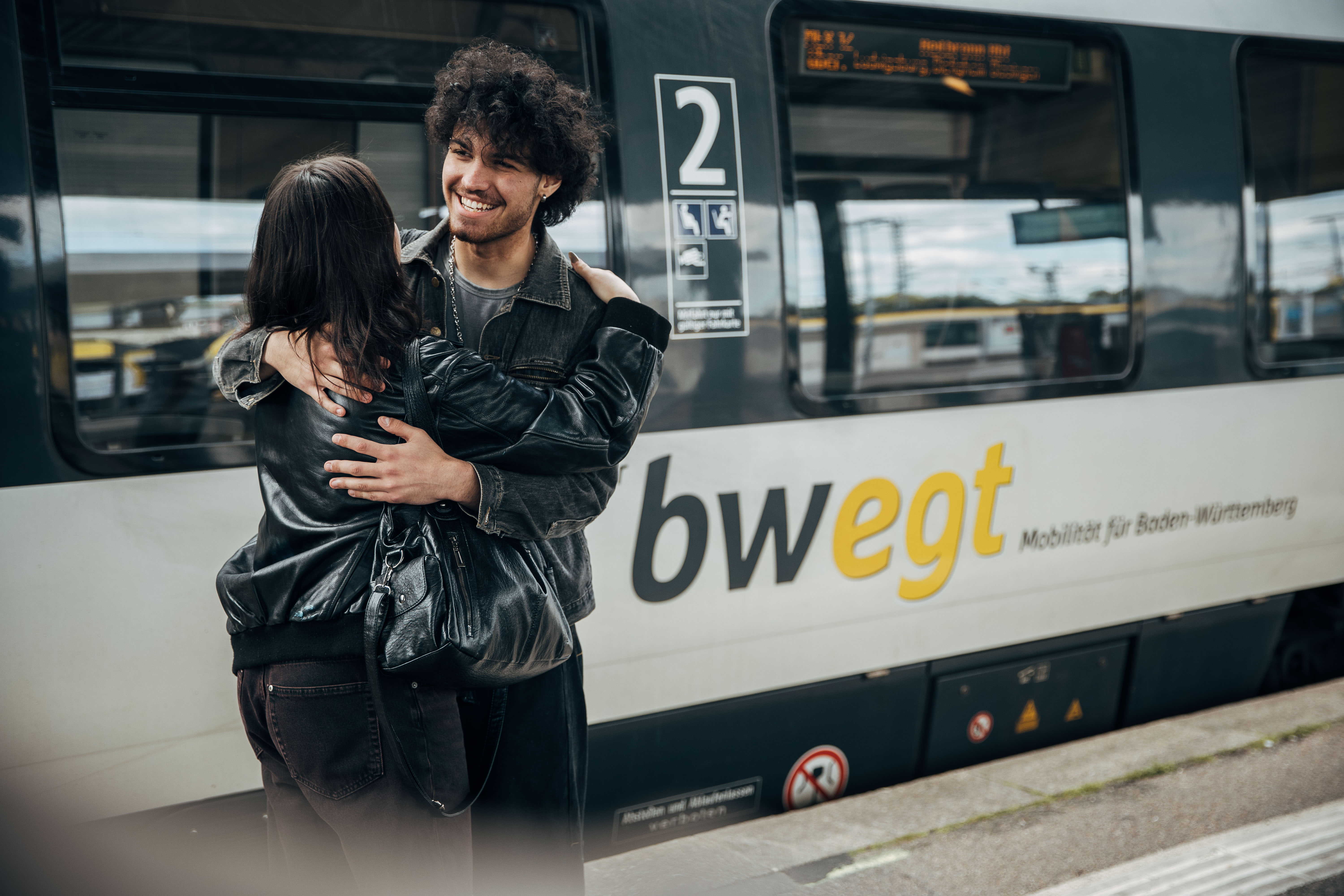 Ein junger Mann mit lockigen dunklen Haaren umarmt eine Frau in schwarzer Lederjacke vor einem stehenden bwegt-Zug. Beide lächeln sich an. Auf dem Zug ist deutlich das bwegt-Logo mit dem Schriftzug „Mobilität für Baden-Württemberg“ sichtbar.