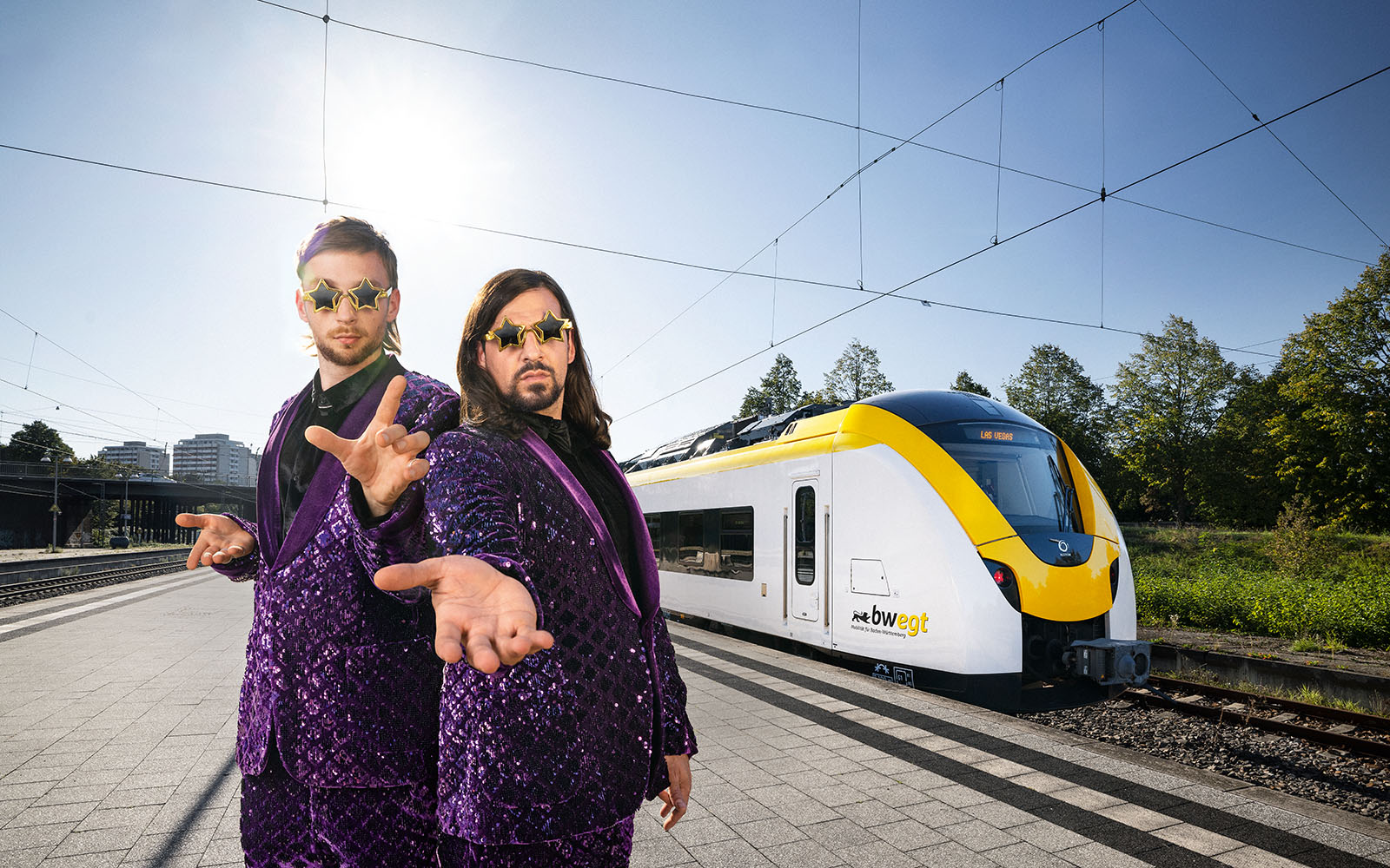 Zwei Zauberer in violetten Anzügen und Sonnenbrillen stehen vor einem bwegt-Zug