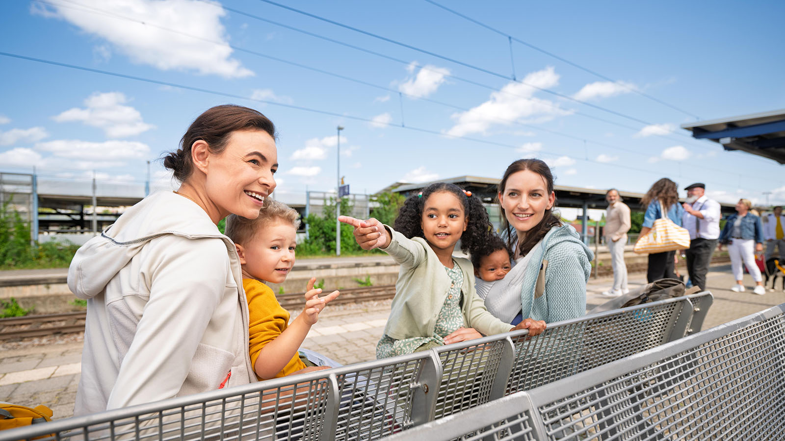 Zwei Mütter sitzen mit ihren drei kleinen Kindern auf einer Bank am Bahnhof
