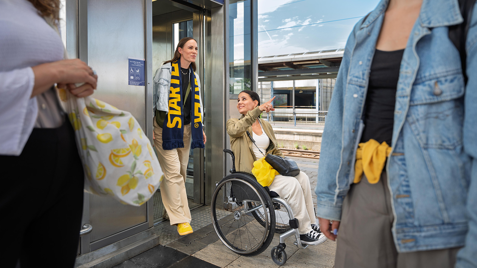 Zwei Frauen, eine davon im Rollstuhl, verlassen einen Aufzug auf einem Bahnsteig